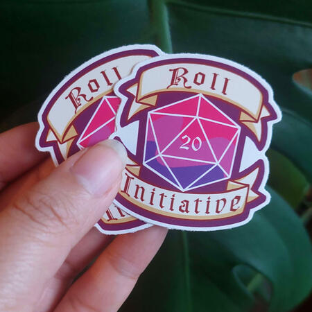 Sticker Two stickers held up that depict a D20 in the bi-flag colour scheme and a banner reading "Roll Initiative"